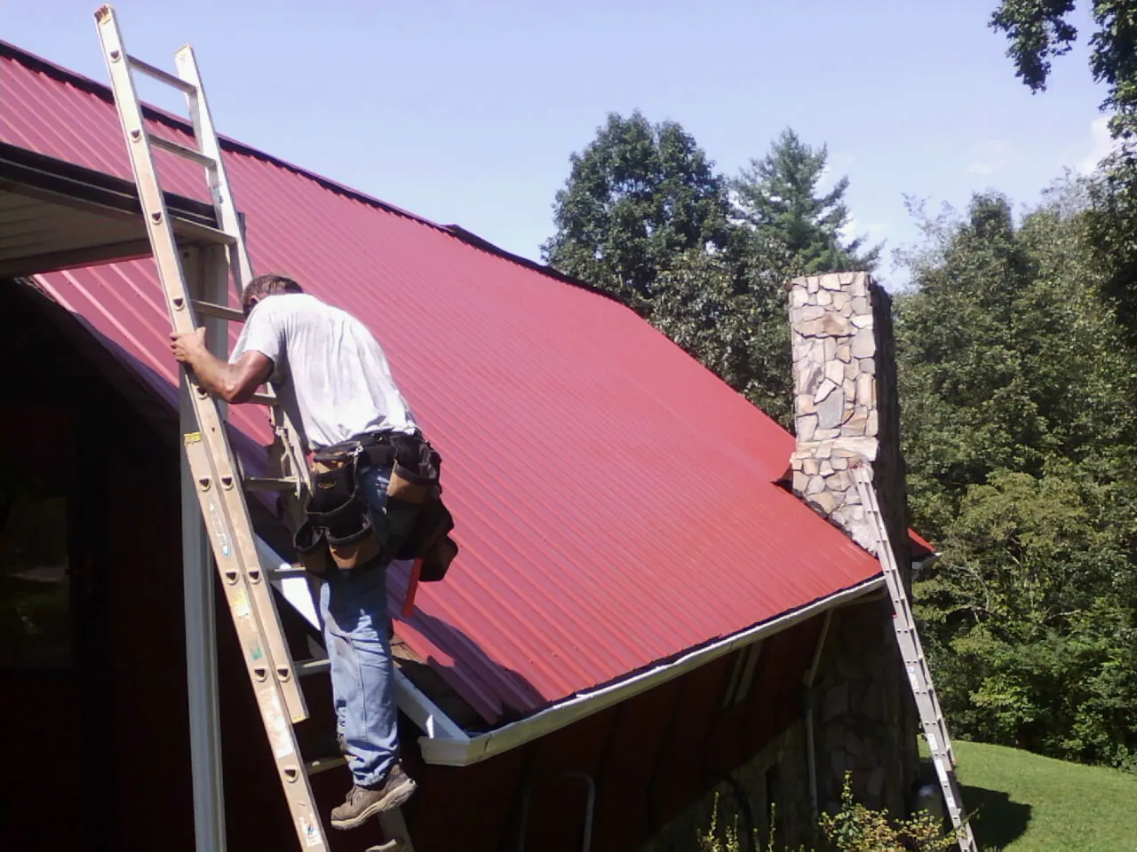 Roofer on a ladder beside a red metal roof and stone chimney — Soffit & Fascia Repair by Roofing Brothers in Carbon Cliff
